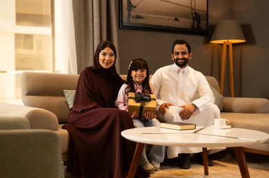 Expressions of happiness and joy, celebrating happy family occasions, upbringing with love and affection, a Saudi Arabian Gulf girl wearing casual attire sitting with her parents in the living room holding a gift box in her hands, the concept of a happy family. Expressions of happiness and joy, celebrating happy family occasions, upbringing with love and affection, a Saudi Arabian Gulf girl wearing casual attire sitting with her parents in the living room holding a gift box in her hands, the concept of a happy family.