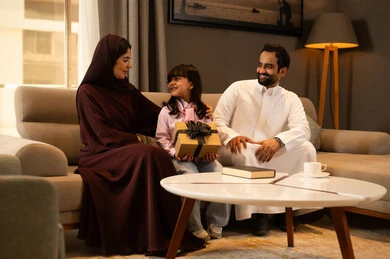 Celebrating happy family occasions, raising children with love and affection, a Saudi Arabian Gulf girl wearing casual attire sits with her parents in the living room holding a gift box in her hands, the concept of a happy family, expressions of happiness and joy. Celebrating happy family occasions, raising children with love and affection, a Saudi Arabian Gulf girl wearing casual attire sits with her parents in the living room holding a gift box in her hands, the concept of a happy family, expressions of happiness and joy.