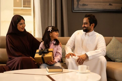 Education with love and affection, celebrating happy family occasions, a Saudi Arabian Gulf girl wearing casual attire sitting with her parents in the living room holding a gift box in her hands, the concept of a happy family, expressions of happiness and joy.