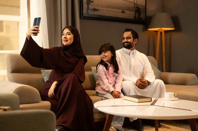 الاجتماع الأسري في غرفة المعيشة وبناء مجتمع سعودي متنامي, التقاط سيلفي عائلي تذكاري عبر الهاتف المحمول,عائلة عربية سعودية خليجية مبتسمة تقوم بتوثيق اللحظات الممتعة بإيماءات السعادة والفرح, فرحة الأبناء بقضاء وقت ممتع مع الأهل