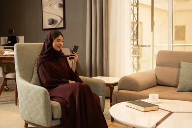 تصفح وسائل التواصل الاجتماعي عبر الجوال، استخدام جهاز تقني حديث, مراه عربيه خليجيه سعودية محجبة ترتدي العباءة تجلس فى غرفه المعيشة تمسك الهاتف المحمول بيدها, تعابير السعادة والسرور