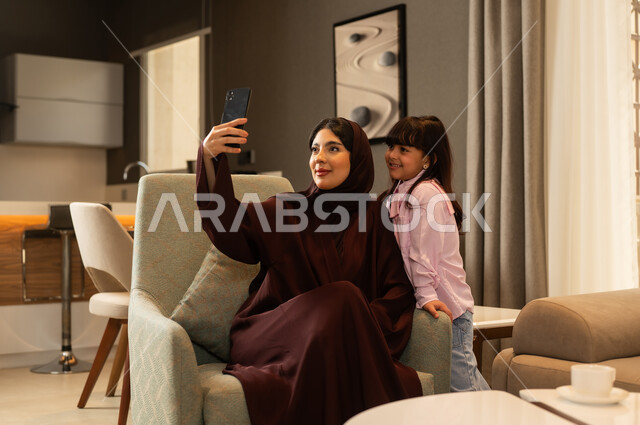 مفهوم الأمومة والطفولة، إجراء مكالمة فيديو، امراة عربيه خليجية سعودية محجبه ترتدي العباءة تجلس فى غرفه المعيشة برفقة ابنتها تقوم بإلتقاط صورة سيلفي تذكارية بإستخدام الهاتف المحمول، رعاية الأم لأطفالها والعناية بهم ، قضاء أوقات ممتعة مع الابناء، الحب والحنان الاسري