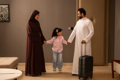 A Saudi Arabian Gulf man wearing a white thobe is holding a travel bag in his hands, standing with his wife and daughter, maintaining family ties, preparing to spend the summer vacation and go on an enjoyable trip, in a family atmosphere filled with love.