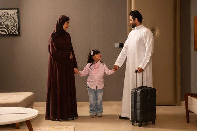 Preparing to spend the summer vacation and go on an enjoyable trip, an Arab Gulf Saudi man wearing a white thobe is holding a travel bag in his hands, standing with his wife and daughter, maintaining family ties, and enjoying a family atmosphere filled with love.