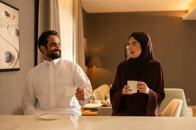 زوجان عربيان خليجيان سعوديان يرتديان الزي التقليدي يجلسان فى غرفه المعيشة يستمتعان بتبادل الحديث ، تناول المشروبات الباردة، نقاشات وحوارات، قضاء أوقات ممتعة، أجواء عائلية سعيدة