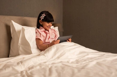 متابعة  حصص الاونلاين عن طريق التابلت ، التعلم عن بعد ، مفهوم التعليم عبر الانترنت ، فتاه عربيه خليجي سعودية ترتدي الزي الكاجوال تجلس على السرير فى غرفه النوم تمسك بيديها الجهاز اللوحي