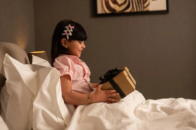 النظر بإيماءات السعادة والسرور، تقديم الهدايا في المناسبات السعيدة، التعبير عن المحبة والتقدير، الاحتفال بأعياد الميلاد, فتاه عربيه سعوديه خليجيه ترتدي الزي الكاجوال تجلس على السرير تمسك بيديها علبة هدية