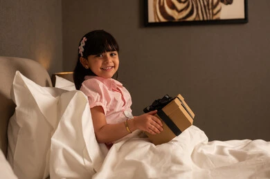 Celebrating birthdays, expressing love and appreciation, giving gifts on happy occasions, a Saudi Arabian Gulf girl wearing casual attire sitting on the bed holding a gift box in her hands, looking with gestures of happiness and joy.