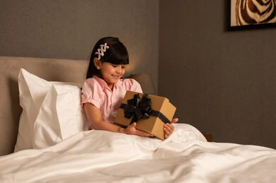 Giving gifts on happy occasions, expressing love and appreciation, celebrating birthdays. An Arab Saudi Gulf girl wearing casual attire sits on the bed holding a gift box in her hands, looking with gestures of happiness and joy.