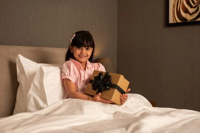 Expressing love and appreciation, celebrating birthdays, giving gifts on happy occasions, a Saudi Arabian Gulf girl wearing casual attire sits on the bed holding a gift box in her hands, looking with gestures of happiness and joy.