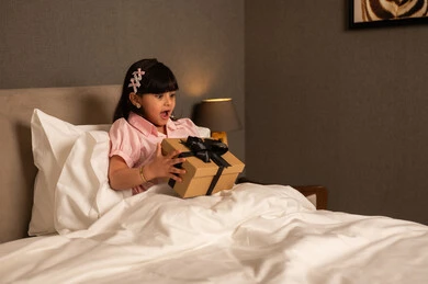 ايماءات المفجاءة و الاندهاش، الاحتفال بأعياد الميلاد, التعبير عن المحبة والتقدير, تقديم الهدايا في المناسبات السعيدة, فتاه عربيه سعوديه خليجيه ترتدي الزي الكاجوال تجلس على السرير تمسك بيديها علبة هدية