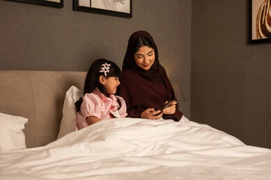 مفهوم الأمومة والطفولة، إجراء مكالمة فيديو، امراة عربيه خليجية سعودية محجبه ترتدي العباءة تجلس فى غرفه النوم برفقة ابنتها تقوم باستخدام الهاتف المحمول، رعاية الأم لأطفالها والعناية بهم ، قضاء أوقات ممتعة مع الابناء، الحب والحنان الاسري