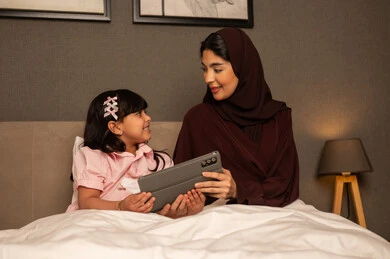 استخدام جهاز تقني حديث، مفهوم التربيه بالحب والحنان، المشاركه بقراءه القصص المفيده مع الأبناء، امراة عربيه خليجية سعودية محجبه ترتدي العباءة تجلس فى غرفه النوم تقرأ الروايات المسليه لابنتها في غرفه النوم عبر جهاز التابلت, استغلال الاوقات في تعليم وتربيه الابناء