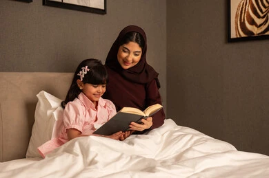 المشاركه بقراءه القصص المفيده مع الأبناء قبل النوم، مفهوم التربيه القائمه على الحب والحنان، قضاء وقت ممتع مع الابنه،  امراة عربيه خليجية سعودية محجبه ترتدي العباءة تجلس فى غرفه النوم تقرأ الروايات المسليه لابنتها