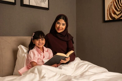 مفهوم التربيه القائمه على الحب والحنان، قضاء وقت ممتع مع الابنه، المشاركه بقراءه القصص المفيده مع الأبناء قبل النوم،  امراة عربيه خليجية سعودية محجبه ترتدي العباءة تجلس فى غرفه النوم تقرأ الروايات المسليه لابنتها