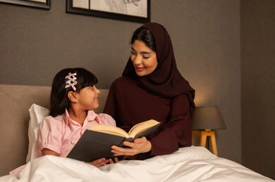 Spending quality time with the daughter, the concept of parenting based on love and affection, sharing the reading of useful stories with the children before bedtime, an Arab Gulf Saudi woman wearing a hijab and an abaya sitting in the bedroom reading entertaining novels to her daughter.