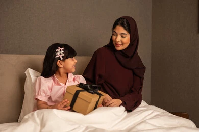 إنشاء الأطفال على الحب والعطاء والتربية الصالحة، الأستمتاع بالأوقات مع الأبناء، فتاة عربيه خليجيه سعودية تجلس فى غرفه النوم تحتفل بعيد الأم وتقدم لوالدتها الهدية، ايماءات تدل على الفرح و السعادة، الإحتفال سويا في الأعياد والمناسبات