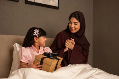 Enjoying time with children, raising kids on love, giving, and good upbringing, a Saudi Arabian Gulf girl sitting in the bedroom celebrating Mother's Day and presenting a gift to her mother, gestures indicating joy and happiness, celebrating together during holidays and occasions.