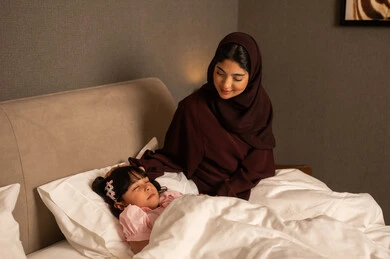 The concept of caring for and paying attention to children, spending happy family times, an Arab Gulf Saudi woman in a hijab wearing an abaya sitting in the bedroom next to her daughter, relaxing and enjoying the calm atmosphere, following a healthy lifestyle.