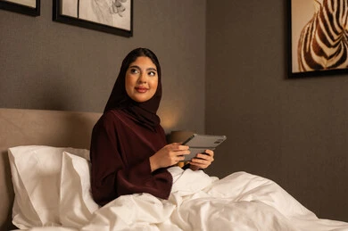 Looking at something, communicating with family and friends, integrating modern technology into daily life, an Arab Gulf Saudi woman wearing a hijab and an abaya sitting on the bed in the bedroom using a tablet, expressions of happiness and joy.
