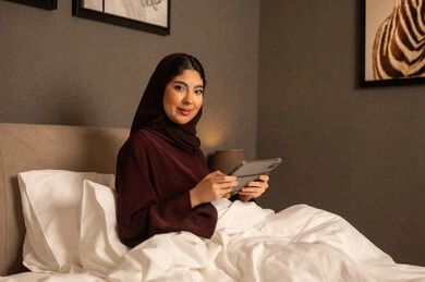 The integration of modern technology into daily life, a Saudi Arabian Gulf woman wearing a hijab and an abaya sits on the bed in the bedroom using a tablet, communicating with family and friends, expressions of happiness and joy.