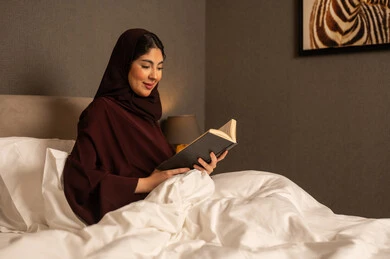 Spending free time on learning and culture, reading with contemplation and focus, an Arab Gulf Saudi woman wearing a hijab and an abaya sits on the bed in the bedroom holding an educational book in her hand.