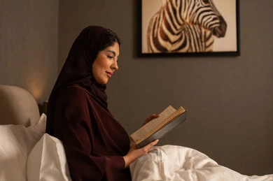 Reading with attention and focus, spending free time on learning and culture, a Gulf Arab Saudi woman wearing a hijab and an abaya sitting on the bed in the bedroom holding an educational book in her hand.