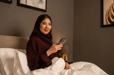 Leisure and entertainment in free time, browsing social media sites, an Arab Gulf Saudi woman wearing a hijab and an abaya sitting on the bed in the bedroom holding a mobile phone, using modern and advanced devices and technologies.
