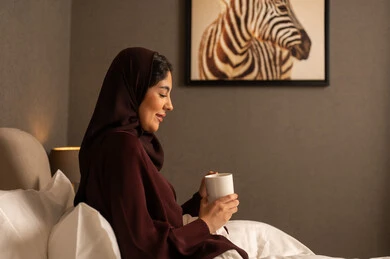 احتساء المشروب المفضل، قضاء وقت ممتع و هادئ، صورة من الجانب لامراه عربيه خليجية سعودية محجبه ترتدي العباءة تجلس على السرير فى غرفه النوم تحمل بيدها فنجان من الشاي، ايماءات السعادة والسرور