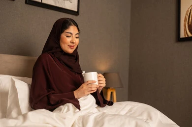 Sitting and enjoying hot drinks and warm winter herbs, spending a pleasant and quiet time, sipping the favorite drink, an Arab Gulf Saudi woman in a hijab wearing an abaya sitting on the bed in the bedroom holding a cup of tea in her hand, gestures of happiness and joy.