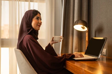 Watching programs and movies, making a video call, an Arab Gulf Saudi woman wearing a hijab and an abaya sitting at the desk working on a laptop, enjoying a hot drink.