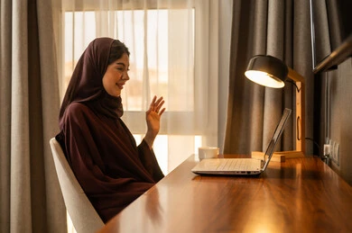 اجراء مكالمة الفيديو،رفع كف اليد بتعابير التحية و السلام، انجاز مهام العمل عن بعد، امراه عربيه خليجية سعودية محجبه ترتدي العباءة تجلس على المكتب تعمل على جهاز الحاسب المحمول ، دمج التكنولوجيا المتطوره فى الحياه اليوميه