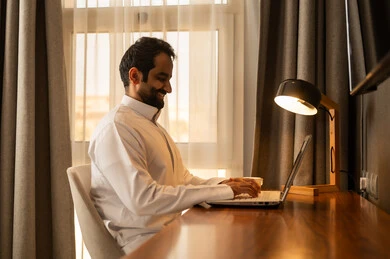 إيماءات التركيز في شاشة الكمبيوتر، استخدام جهاز تقني حديث لإنجاز المهام والمشاريع, تصفح مواقع التواصل الاجتماعي والعمل عن بعد, رجل عربي خليجي سعودي يرتدي الثوب الابيض يجلس على المكتب يستخدم الحاسوب المحمول
