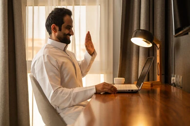 رفع اليد بتعابير التحية و السلام، اجراء مكالمة فيديو اونلاين، تصفح التطبيقات والبرامج الالكترونية عبر الابتوب, استخدام الأجهزة التقنية الحديثة والمتطورة, إدراة الأعمال عن بعد, رجل عربي خليجي سعودي يرتدي الثوب الابيض يجلس على المكتب يستخدم الحاسوب المحمول