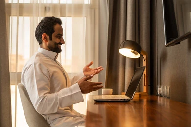 Making an online video call, browsing applications and electronic programs via the laptop, using modern and advanced technological devices, managing business remotely, an Arab Gulf Saudi man wearing a white thobe sitting at the desk using a laptop.