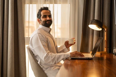 احتساء المشروب الساخن، تصفح التطبيقات والبرامج الالكترونية عبر الابتوب, استخدام الأجهزة التقنية الحديثة والمتطورة, إدراة الأعمال عن بعد, رجل عربي خليجي سعودي يرتدي الثوب الابيض يجلس على المكتب يستخدم الحاسوب المحمول