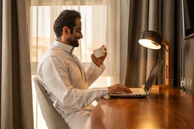 Enjoying a hot drink, browsing applications and electronic programs on the laptop, using modern and advanced technological devices, managing business remotely, an Arab Gulf Saudi man wearing a white thobe sitting at the desk using a laptop.