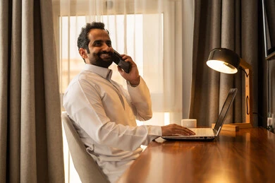 Browsing applications and electronic programs via the laptop, making a phone call via the mobile, using modern and advanced technological devices, managing business remotely, an Arab Gulf Saudi man wearing a white thobe sitting at the desk using a laptop.