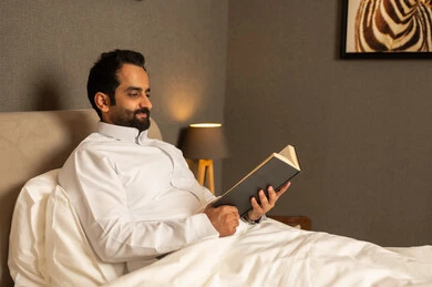 Enjoying reading novels and stories, reading books attentively, the culture of self-learning, enjoying reading novels and stories, practicing the hobby of reading, an Arab Gulf Saudi man wearing a white thobe sitting on the bed in the bedroom holding an educational book in his hand.