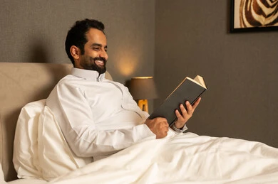 قراءة الكتب بتمعن، ثقافة التعلم الذاتي، الاستمتاع بقراءة الروايات والقصص، ممارسة هواية المطالعة ، رجل عربي خليجي سعودي يرتدي الثوب الابيض يجلس على السرير فى غرفه النوم يحمل بيده كتاب تثقيفي ، الاستمتاع بقراءة الروايات والقصص