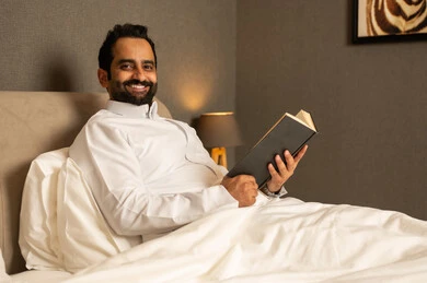 The culture of self-learning, enjoying reading novels and stories, practicing the hobby of reading, reading books attentively, an Arab Gulf Saudi man wearing a white thobe sitting on the bed in the bedroom holding an educational book, enjoying reading novels and stories.
