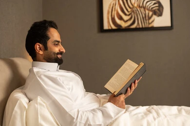 Enjoying reading novels and stories, practicing the hobby of reading, the culture of self-learning, reading books attentively, an Arab Gulf Saudi man wearing a white thobe sitting on the bed in the bedroom holding an educational book, enjoying reading novels and stories.