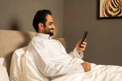 Browsing social media, enjoyment and entertainment using modern technology, a Gulf Arab Saudi man wearing a white thobe sits on the bed in the bedroom holding a mobile phone in his hands, gestures of happiness and joy.