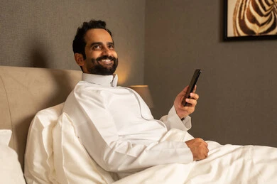 المتعة والتسلية باستخدام التقنية الحديثة،تصفح مواقع التواصل الاجتماعي، رجل عربي خليجي سعودي يرتدي الثوب الابيض يجلس على السرير فى غرفه النوم يمسك بيديه الجوال ، ايماءات السعادة و السرور