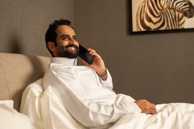 Talking and making a call on a mobile phone, an Arab Gulf Saudi man wearing a white thobe is sitting on the bed in the bedroom holding the mobile phone in his hands, with gestures of happiness and joy.