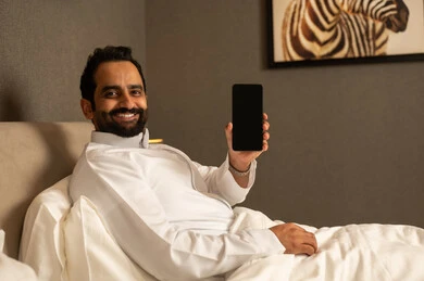 A blank black screen is displayed on the mobile phone, an Arab Gulf Saudi man wearing a white thobe is sitting on the bed in the bedroom holding the phone in his hands, showing gestures of happiness and joy. A blank black screen is displayed on the mobile phone, an Arab Gulf Saudi man wearing a white thobe is sitting on the bed in the bedroom holding the phone in his hands, showing gestures of happiness and joy.