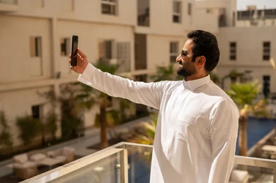 Video chat through social media applications, taking commemorative selfie photos via mobile, using modern devices and technologies, an Arab Gulf Saudi man wearing a white thobe and sunglasses holding a mobile phone in his hands, having a good time outdoors, expressions of happiness and joy.