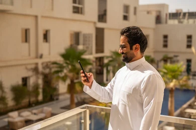 Chatting and communicating with friends with gestures of happiness and joy, using modern devices and technologies, an Arab Gulf Saudi man wearing a white thobe and sunglasses holding a mobile phone in his hands browsing the internet, spending enjoyable time outdoors.