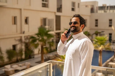 Expressions of happiness and joy, making a phone call via mobile, an Arab Gulf Saudi man wearing a white thobe and sunglasses holding a mobile phone in his hands, spending enjoyable time outdoors, talking with family and friends.