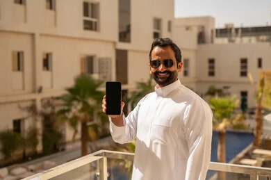 A black screen is displayed on the mobile phone, a Gulf Arab Saudi man wearing a white thobe and sunglasses is holding a mobile phone in his hands, enjoying time outdoors, with expressions of happiness and joy.